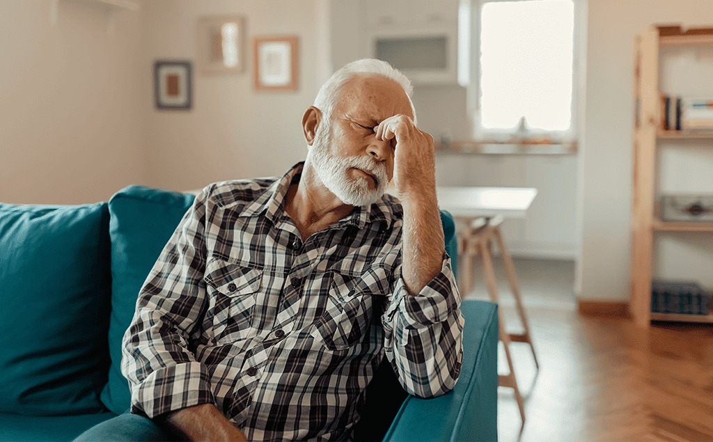 An elderly man sitting on a couch at home holding his forehead looking tired